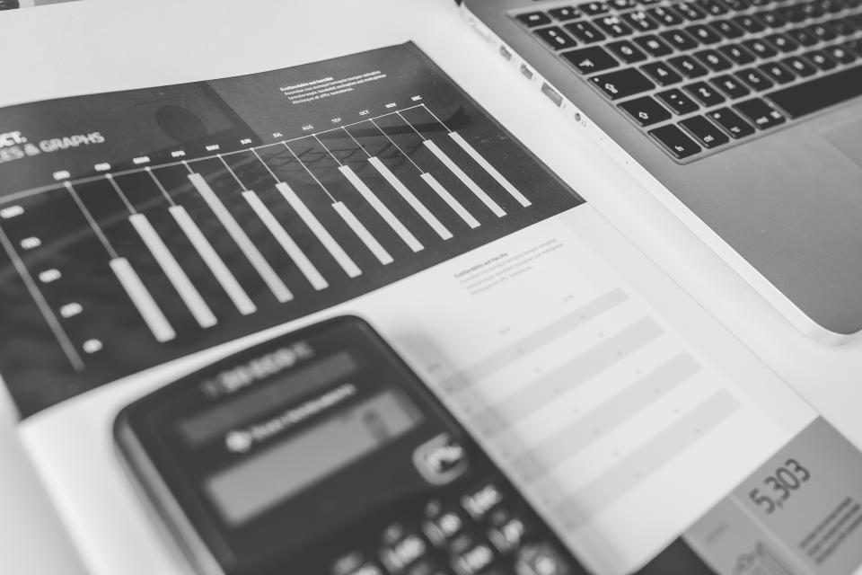 Un foglio con tabelle e numeri accanto a un computer portatile, in bianco e nero